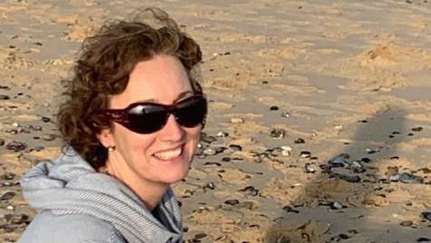 Mrs Cain smiles at the camera as she sits on a beach as the sun sets. She wears sunglasses a grey hoodie.