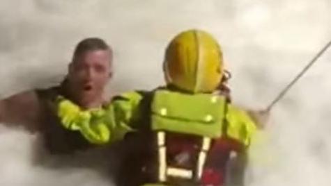 Still from a video of rescue showing firefighter attached to a line holding onto swimmer in rough seas