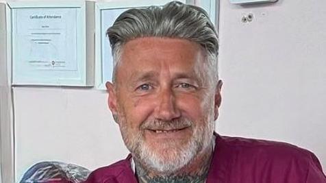 Sean Scott has a grey beard, grey hair and a tattoo on his neck. He's wearing a maroon top and sitting in a room in front of a couple of framed certificates. 