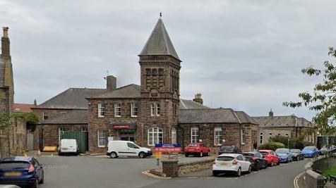 View of a Victorian hospital building. A square bell tower stands to the left of a two-story entrance, with a single story wing to the side, and further building to the rear. Cars are parked on a forecourt to the front, with more parked on a street to the side.