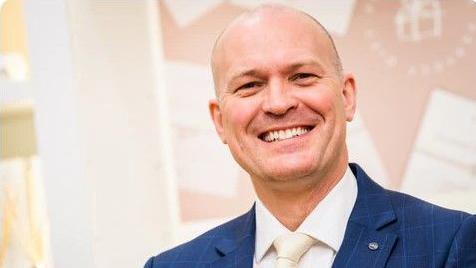 A smiling man wearing a blue suit and cream coloured tie. 