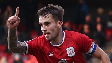 Luke Offord celebrates scoring for Crewe by holding his right arm in the arm and pointing towards the sky with his index finger.