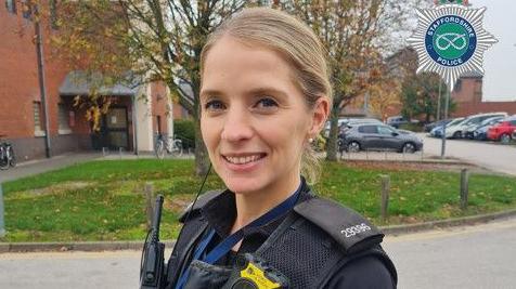 A woman in a police uniform. She has blonde hair tied back into a ponytail and is standing outside in front of a patch of grass and red brick building. 