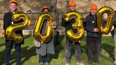 Four people standing outside the Queen Elizabeth Hospital in King's Lynn. They are wearing orange hard hats. Each protester is holding a large gold balloon. The balloons spell out 2030.