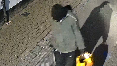 The image shows a person whose face is obscured, carrying a ladder and a yellow bag and walking along a street. It is dark.