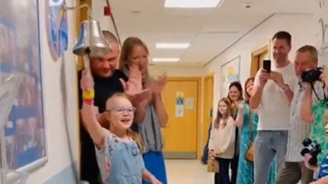 Young girl smiling as she rings a bell while people gather around her smiling and clapping