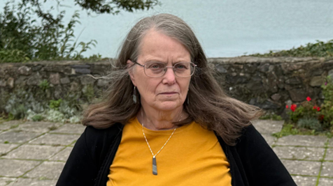Morfudd Mill is standing on a paved area with trees and the sea in the background. She is wearing a mustard yellow top, black cardigan and green pendant. She has long grey and brown hair.