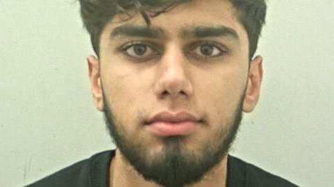 A mugshot of a young man with dark hair and a short beard