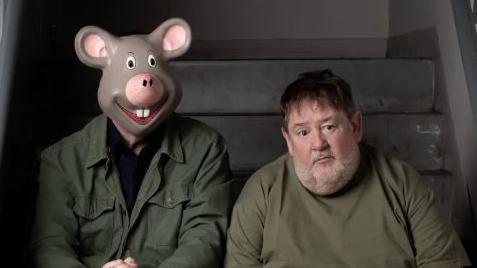 Johnny Vegas sits glumly next to a tall man wearing a mask of a cartoon mouse's head over his face. Both are wearing dark green tops and are sitting on the steps of a staircase.