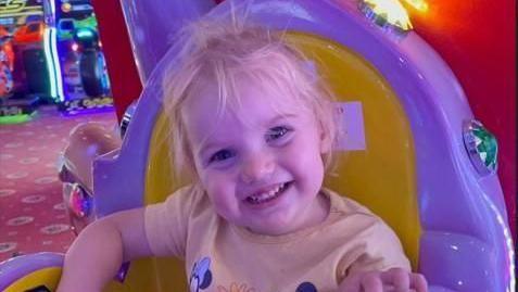 A little girl, Isabella Wheildon, is wearing a yellow top and sitting in a yellow and purple chair at an amusements arcade. She has blonde hair and is smiling at the camera.