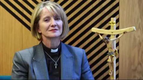 The Bishop of Warrington - a woman with bobbed blonde hair wearing a grey jacket, black shirt and dog collar, standing next to a crucifix 