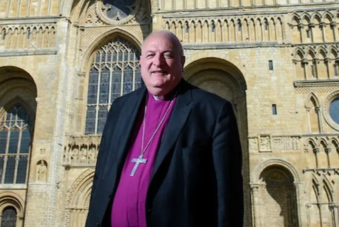 Bishop Conway, in religious attire with a crucifix around his neck, stands in front of a cathedral building. He is smiling.