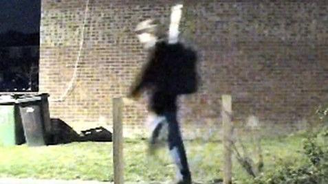 Grainy nighttime CCTV images of a man in a cap walking in front of a brick building with some bins in front of it. 
