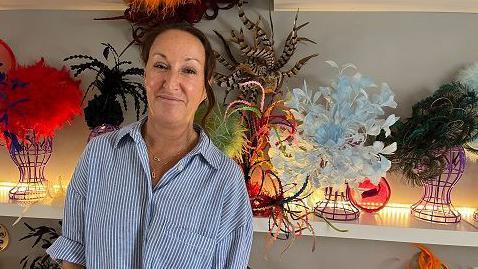 Carrie Jenkinson looks into camera with a selection of colourful hats and fascinators behind her