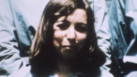 A blurred photograph of Lesley Whittle. She has shoulder-length brown hair and is smiling at the camera. Behind her are people wearing blue tops but we cannot see their hands or faces due to how close they are to Lesley.