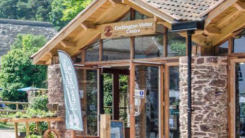 The entrance of the building. It is a brick building and it has glass doors and windows. There are trees around it.