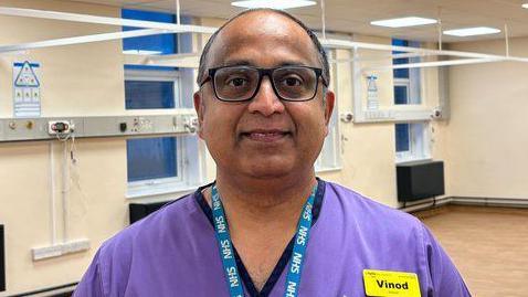 Vinod Mani, a hospital matron, stands in an empty room, which is painted cream with white windows and black radiators. It has light wooden flooring. Mr Mani is middle aged, and is wearing glasses, purple scrubs, a yellow name badge, and a blue NHS lanyard.