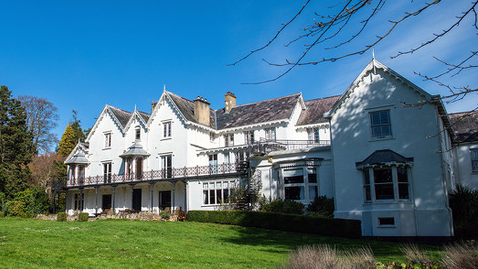 The three-storey white frontage of the Richmond Hotel on a sunny day. There are tall trees to the left of the hotel and grass in front.