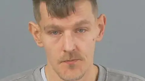 A mugshot of Ross Neiland, who is wearing a grey jumper in front of a grey background. He has a goatee beard and moustache and short brown hair. He is looking at the camera.