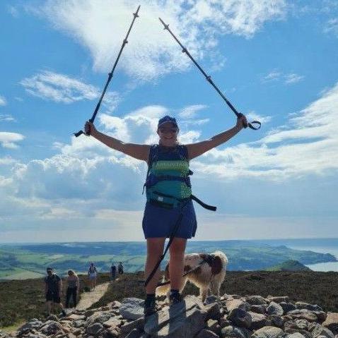 Rachel at the top of Great Hangman - standing on a rock cairn with her dog. It's blue sky and hills behind. People are walking along the path at the top.