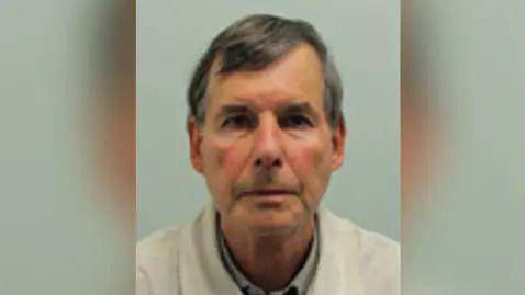 A police mugshot of a man with grey head and wearing a white jacket.