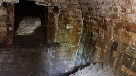 The interior of a curved brick built building with soil in the foreground and an entrance to another room in the background