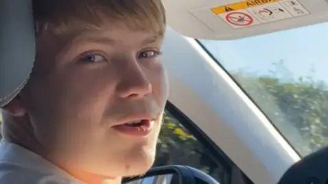 A man with blond hair sat in the front passenger seat of a car, looking directly at the camera.