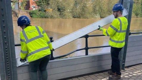 Two people are dressed in bright yellow hi-vis jackets, and are wearing blue helmets. Their jackets say "Environment Agency". They are holding a long slat of a flood barrier, and are inserting it between two metal barriers. In the background is brown river water and green trees.