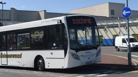 Single-decker white bus at a junction outside a hospital entrance.  There is a small white van in the background, and a blue road sign directing traffic.