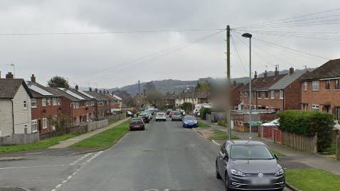 A residential road with houses and cars on either side and hills pictured in the distance. 