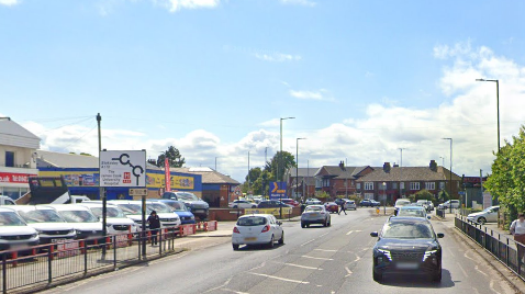 The approach to a roundabout with a Kwikfit garage to the left and railings to the right. Cars are travelling in both directions.