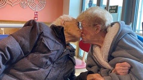 Maureen and Harry are sitting together and kissing. They have white hair and are dressed in warm clothing