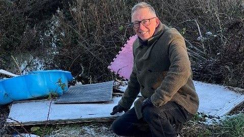 Phillip is wearing a light green jacket and black gloves. He has grey hair and dark-rimmed glasses and is bending down, smiling at the camera at the site of fly-tipped rubbish. Included in the rubbish is a plastic blue children's bathing pool and pink mat