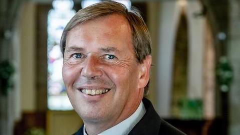 A smiling vicar dressed in a clerical collar and shirt and jacket is photographed in his church.
