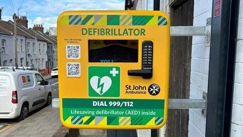 A close-up of a St John Ambulance defibrillator machine at Luton Town Football Club. The device is in a yellow and green box with the word "Defibrillator" written on it. It on a wall outside the ground mounted on two metal bars. There are terraced houses and cars visible behind it.