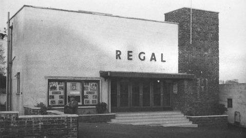 Black and white photograph of a cinema called the Regal with a man resting a bag on a wall outside
