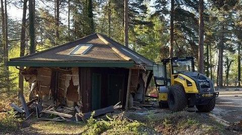 A wooden toilet block with a shallow pyramid shaped roof at the edge of a car park in a forest setting. Its sides are smashed and wrecked. A construction vehicle is next to it with smashed windows.