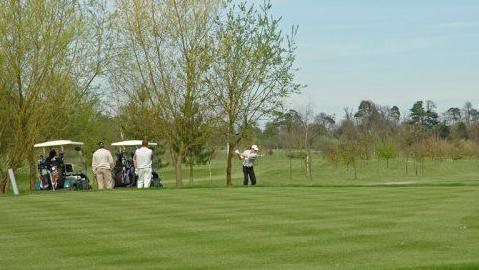 A golfer wearing a white hat, pale jumper and dark trousers, playing a drive at a golf course. A number of other people are standing behind the officer and watching.