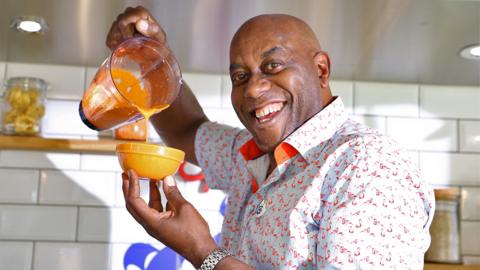 A grinning Ainsley Harriott pours his bright orange red pepper rouille sauce into a glass dish.