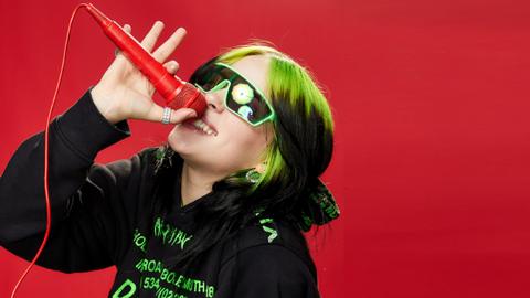 Billie Eilish is holding a microphone in the air, she is wearing sunglasses and is smiling.