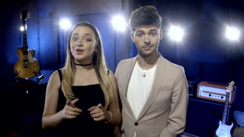 Girl with long hair and wearing a black dress pointing at the camera with both hands. A young man wearing a brown coat is standing next to her.