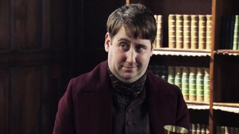 Thomas Edison from Horrible Histories holding a Victorian telephone in his hands. He has a cunning look on his face.