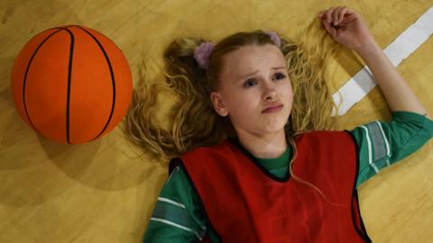 A young blonde girl is looking confused lying on the floor next to a basketball