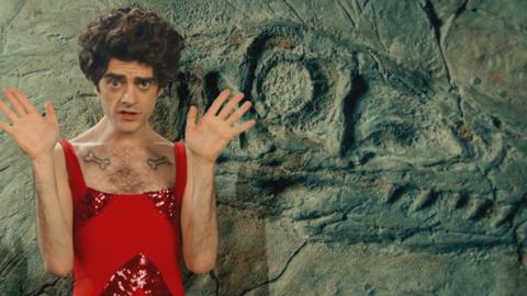 A man in a red jumpsuit poses in front of a dinosaur fossil