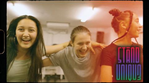 Three girls are smiling and laughing whilst holding each other's shoulders