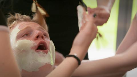 A boy cries out as girls attack him with a face mask and make up.