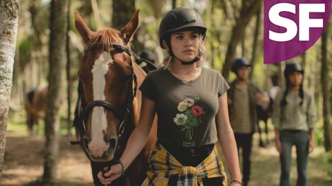 A young girl wearing a riding helmet, has her shirt tied around her waist. She is holding a large brown horse by it's reign next to her as she looks confusingly into the distance