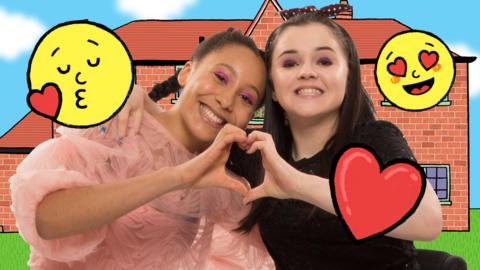 Two young girls are sat side by side smiling to camera, they are creating a love heart with their hands.