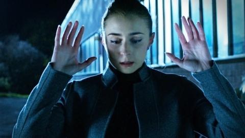 A girl with a dark grey jacket and her eyes cast down puts her hands up in the air in a sign of surrender.