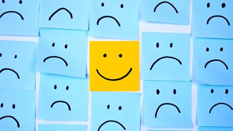 A wall covered in blue post it notes with sad faces on them. In the centre, sits a yellow post-it note with a smiley face.